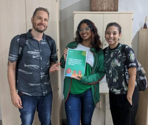 Foto com participantes do projeto do guia grêmios: um homem branco na esquerda e duas estudantes negras à direita, com a do meio segurando a imagem do Guia Grêmios